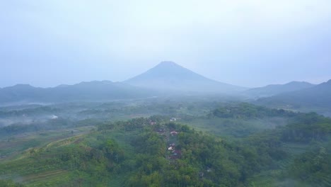 Paisaje-Verde-De-Sobrevuelo-Aéreo-Lento-Con-Un-Pequeño-Pueblo-Y-Montañas-Nubladas-En-Indonesia