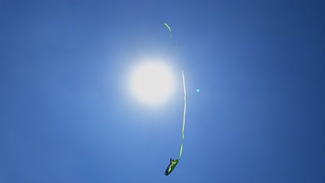 Unusual-perspective-of-flying-green-toy-kite-close-to-shining-sun-in-clear-blue-sky