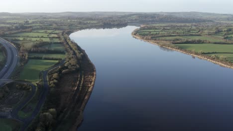 Motorway-and-green-pastures-abut-smooth-reflective-river-water,-aerial