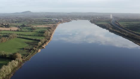 Aerial-flies-over-wide,-glassy-smooth-river-amid-rural-agriculture