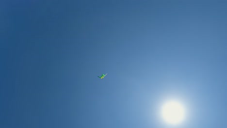 Low-angle-view-of-flying-green-toy-kite-close-to-shining-sun-in-clear-blue-sky
