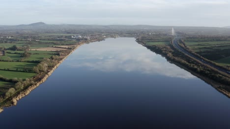 Aerial:-Glassy-smooth-river-reflects-sky-and-clouds,-distant-bridge