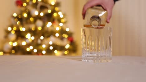 Hand,-Die-Whiskey-In-Kristallglas-Auf-Einem-Küchentisch-Gießt-Und-Vor-Einem-Beleuchteten-Und-Geschmückten-Weihnachtsbaum-Zu-Hause-Feiert,-Zeitlupe