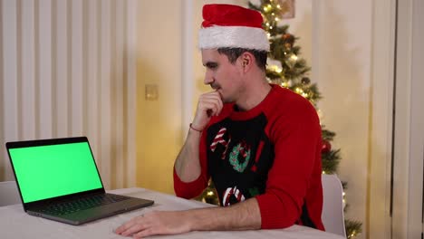 Ein-Festlicher-Mann-Vor-Einem-Weihnachtsbaum,-Der-Tief-In-Gedanken-Versunken-Auf-Einen-Greenscreen-Laptop-Starrt-Und-Besorgt-über-Das-Aussieht,-Was-Er-In-Dem-Video-Sieht