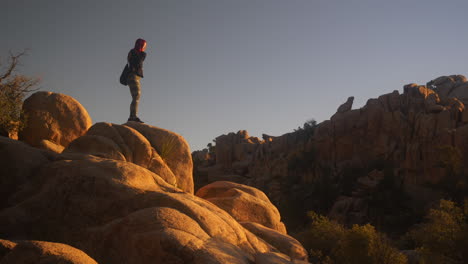 Wide-Shot-Red-Haired-Girl-Climbs-on-top-of-Boulder-and-poses-at-Joshua-Tree,-California