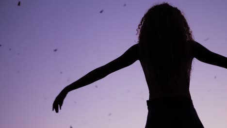 A-woman-raising-her-hands-to-the-sky-and-nature,-dancing-under-the-purple-night-sky-with-bats-flying-high---Slow-motion