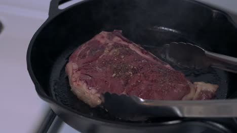 Dinner-steak-cooked-on-a-cast-iron-skillet-ready-to-flip-over-with-stainless-steel-tongs
