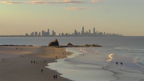 Menschen,-Die-Bei-Sonnenuntergang-Am-Strand-Von-Currumbin-Spazieren-Gehen-Und-Surfen---Currumbin-Alley-Und-Surfers-Paradise-Hochhäuser---Blick-Vom-Elefant-Rock-Lookout---Gold-Coast,-Qld,-Australien