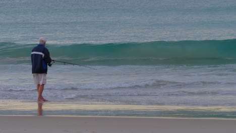 An-Old-Man-Catching-Fish-With-A-Fishing-Rod-At-The-Coast---Wave-Rolling-Ashore-In-Currumbin-Beach---Fishing-In-Gold-Coast,-QLD,-Australia---wide-shot