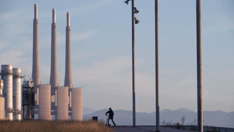 Man-contemplating-industrial-landscape-rides-off-on-electric-scooter-at-sunset