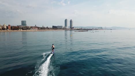 Aerial-follow-shot-of-fast-surfer-riding-electric-surfboard-in-front-of-iconic-Barcelona-skyline-on-a-sunny-day