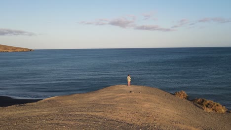 Aerial-drone-shot-of-a-woman-standing-on-a-cliff-in-front-of-the-ocean,-Canary-Islands,-Fuerteventura