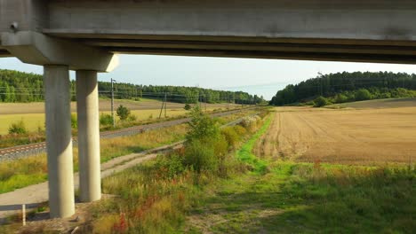 Low-altitude-aerial-drone-view-moving-backwards-following-on-the-side-of-electrified-railroad-tracks-with-forests-and-fields-on-both-sides,-passing-under-road-bridge
