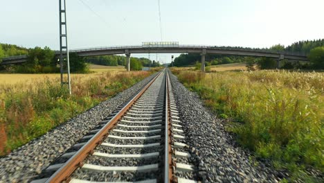 Slow-speed-aerial-drone-view-at-low-altitude-moving-forward-following-straight-railroad-tracks-with-bridge-ahead-and-agricultural-fields-on-both-sides,-simulating-train-ride-in-summer-time