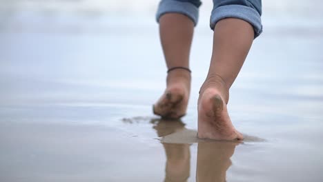 Following-an-undefined-baby-feet-walking-on-beach-with-his-parents-in-afternoon-with-sea-waves-splashing-his-barefoot-legs,-SLOW-MOTION
