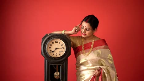 Kolkata:-Traditionell-Gekleidete-Indische-Frau-In-Rot-weißem-Saree-Mit-Einer-Traditionellen-Vintage-Uhr,-Die-In-Einem-Studio-Mit-Rotem-Hintergrund-Denkt