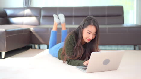 Joven-Mujer-Feliz-Tendida-En-El-Suelo-De-La-Sala-De-Estar-Y-Escribiendo-En-Un-Ordenador-Portátil-Con-Una-Sonrisa-En-La-Cara