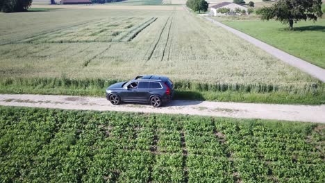 A-Volvo-xc-90SUV-car-drives-slowly-on-a-Small-bumpy-road-inbetween-agriculture-field-after-an-intersection-between-two-roads,-Swiss-countryside,-Vaud,-drone-tracking-aerial-view
