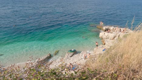 Tourists-in-Turquoise-rocky-beach-in-Kassiopi-coastline,-Panning-shot-from-above,-Corfu-Island