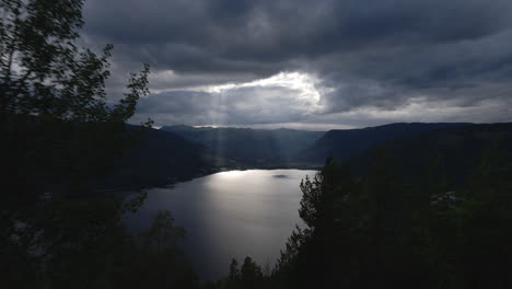 Aerial-shot-of-lake-in-between-the-mountains-with-sunrays-coming-out-from-the-clouds