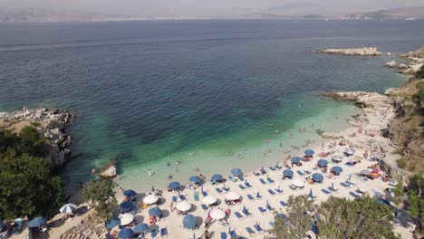 Schöner-Griechischer-Kiesstrand-Mit-Blauen-Sonnenschirmen-Und-Türkisfarbenem-Wasser,-Insel-Korfu