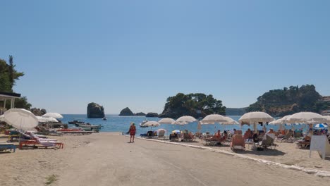 Slow-panning-shot-of-tourists-relaxing-under-parasols-on-the-stunning-beach-at-Parga,-Greece