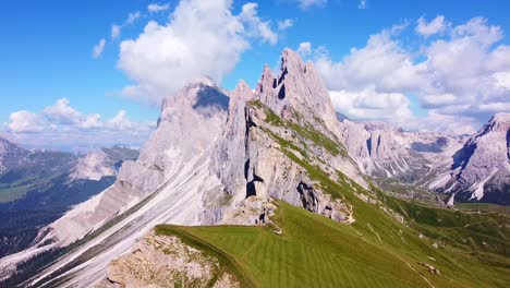 Aerial-view-with-parallax-of-the-peaks-of-Seceda-with-green-pastures,-and-hiking-trails-in-the-foreground,-and-mountain-peaks-in-the-background-in-the-Italian-Dolomites-in-South-Tyrol,-Italy