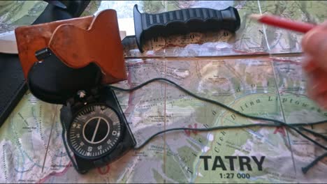 Hitchiker-preparing-to-hike-on-trail-in-mountains-analyzing-the-map-with-a-compass,-pencil-and-knife,-pov,-first-perspective