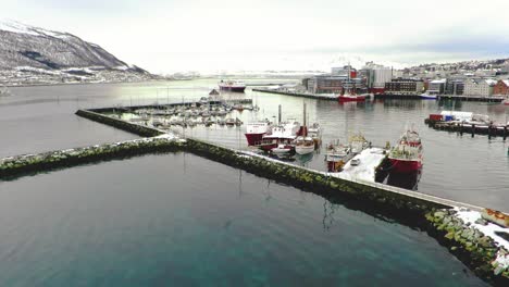 Port-with-ships-and-boats-in-northern-coastal-city-in-winter-with-mountains-and-houses-covered-in-snow-in-Tromso-Norway