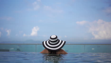 Eine-Frau-Mit-Einem-Schwarz-weiß-Gestreiften-Hut,-Die-Sich-In-Einem-Pool-Entspannt,-Während-Sie-Auf-Das-Meer-Blickt