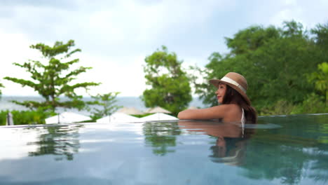 A-young-Asian-woman-wearing-a-brown-hat-leans-over-an-infinity-pool's-edge-while-enjoying-the-outdoor-view-of-a-resort