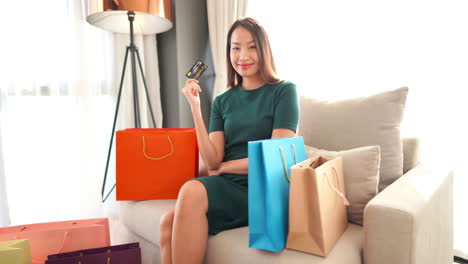 confident-woman-in-a-green-dress-smiling-as-she-holds-her-credit-card-up-while-sitting-in-a-chair-surrounded-by-brightly-colored-designer-shopping-bags