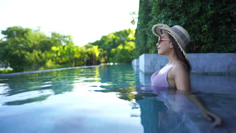 Side-view-of-an-Asian-woman-sitting-in-a-resort-infinity-pool-with-hat