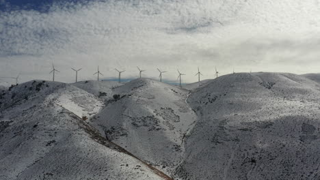 Wind-turbines-spin-on-top-of-snow-covered-mountains-with-clouds,-Aerial