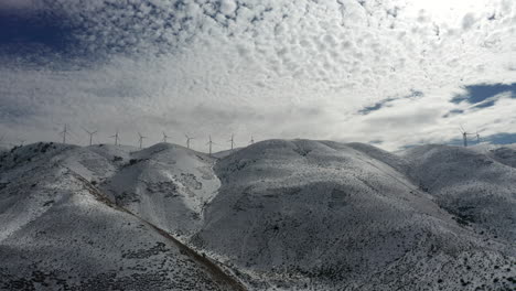Flying-sideways-by-snowy-foothill-mountains-with-row-of-wind-turbines-spinning