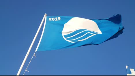 Foundation-for-Environmental-Education-Blue-Flag,-flying-in-a-stiff-wind-over-D'Amour-Beach,-Sidari-Corfu