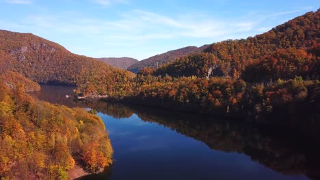 Forward-drone-view-of-lake-Tarnita,-Romania,-surrounded-by-colorful-autumn-trees