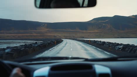 Vista-Del-Coche-Del-Tablero-De-Instrumentos-De-La-Conducción-En-El-Puente-De-La-Carretera-Mojada-Salvaje-De-Islandia-Con-Montañas-En-La-Distancia