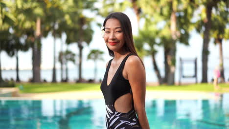 Hermosa-Y-Tímida-Chica-Asiática-Con-Traje-De-Baño-Negro-Sonriendo-A-La-Cámara,-Fondo-Borroso-De-La-Piscina