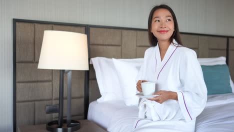 Smiling-Asian-woman-in-white-bathrobe-drinking-tea-while-sitting-on-hotel-bed