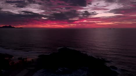 Aerial-ascend-over-Arpoador-rock-dark-in-the-foreground-with-the-cloudy-sky-coloured-red-and-magenta-from-the-sun-still-beneath-the-horizon-before-sunrise