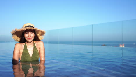 Pretty-fit-Asian-woman-in-a-green-bathing-suit-and-floppy-straw-sun-hat-relaxes-in-a-resort-luxury-pool
