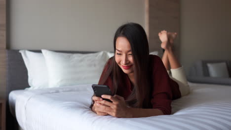 Joyous-asian-lady-texting-on-a-hotel-bed-with-a-broad-smile