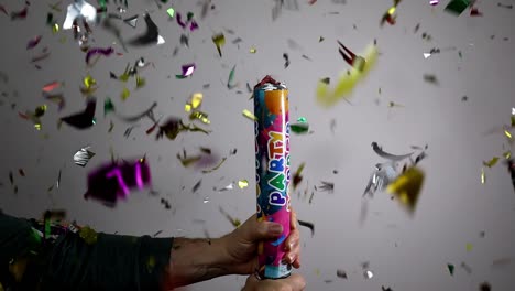 Man-holding-confetti-cannon-twists-tube-and-shiny-colorful-pieces-of-paper-are-launched-into-the-air-then-float-through-the-scene
