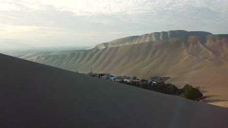 Aerial,-tracking,-drone-shot,-of-a-man-and-woman-sitting-on-a-dune,-looking-at-the-Huacachina-oasis-town,-at-sunset,-on-a-sunny-evening,-in-southwestern-Peru