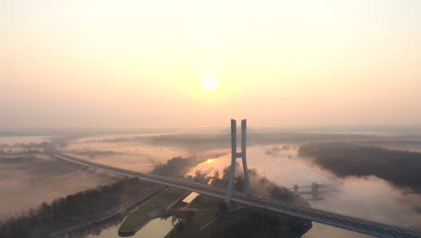 Aerial-shot-of-a-modern-cable-road-bridge,-over-a-wide-calm-river,-during-a-foggy-sunrise