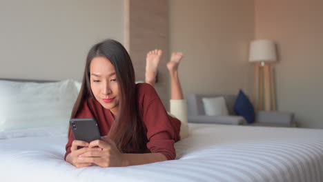 Smiling-asian-girl-lying-on-the-bed-and-texting-with-her-friends