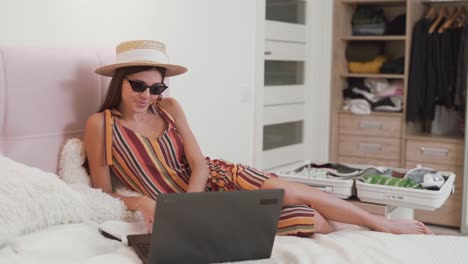 beautiful-girl-in-a-card-in-bed-chooses-on-a-laptop-where-to-fly-on-vacation,-on-the-background-of-a-collected-suitcase