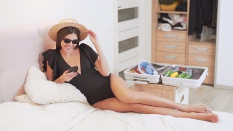 beautiful-girl-in-a-bathing-suit-lying-on-the-bed-chooses-on-the-phone-where-to-fly-on-vacation,-a-suitcase-is-collected-on-the-background