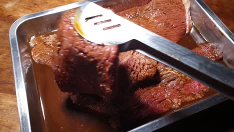 Barbeque-bbq-Grill-Tongs-turning-over-tenderized-and-marinated-beef-steak-in-sauce-inside-a-stainless-steel-pan-before-frying-and-grilling